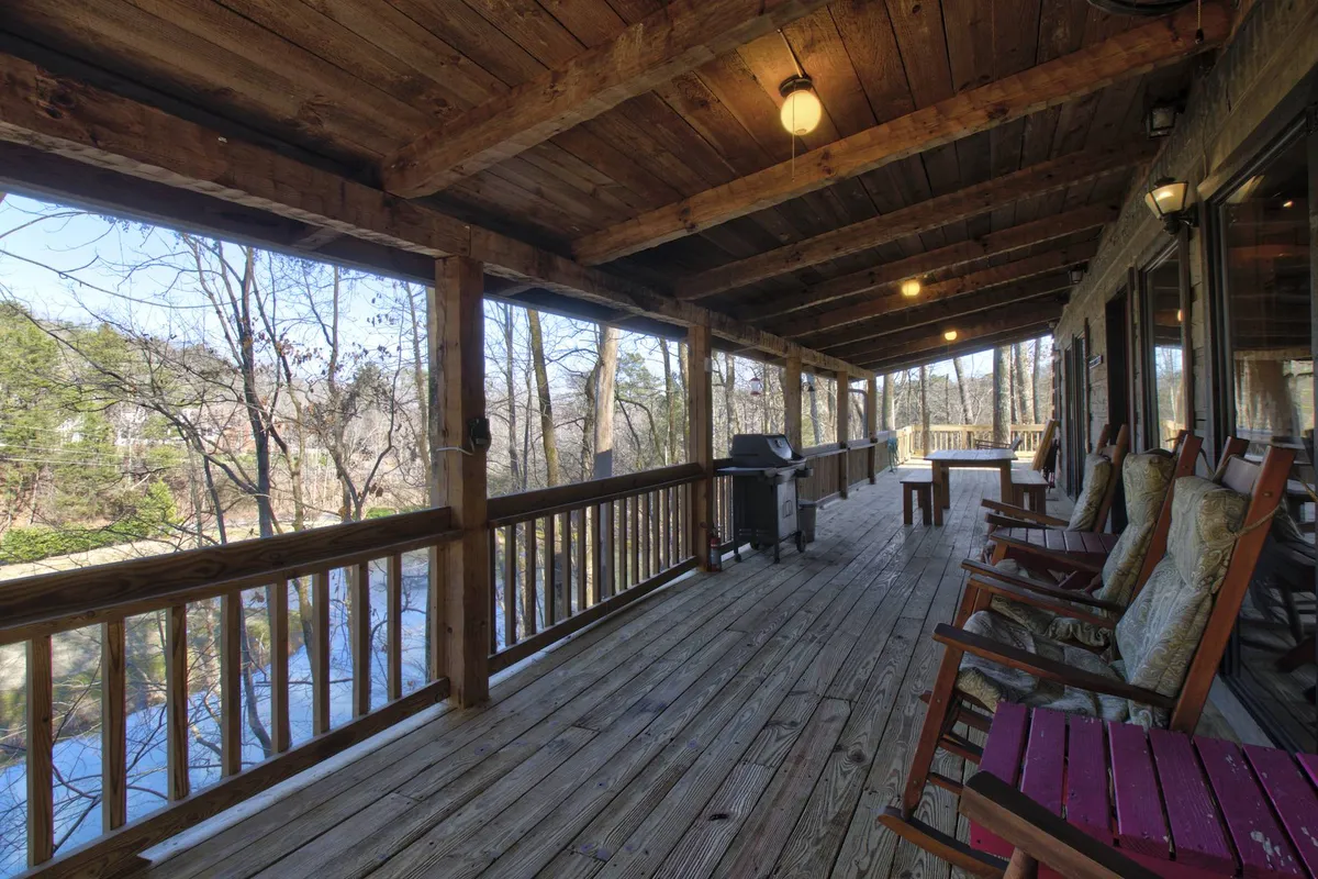 A covered deck with wooden railings overlooks a lush, tree-filled landscape. Rocking chairs are positioned to take in the view, with a grill ready for outdoor cooking. The deck offers glimpses of a pond or water feature through the trees, and the wooden floor adds a rustic touch.