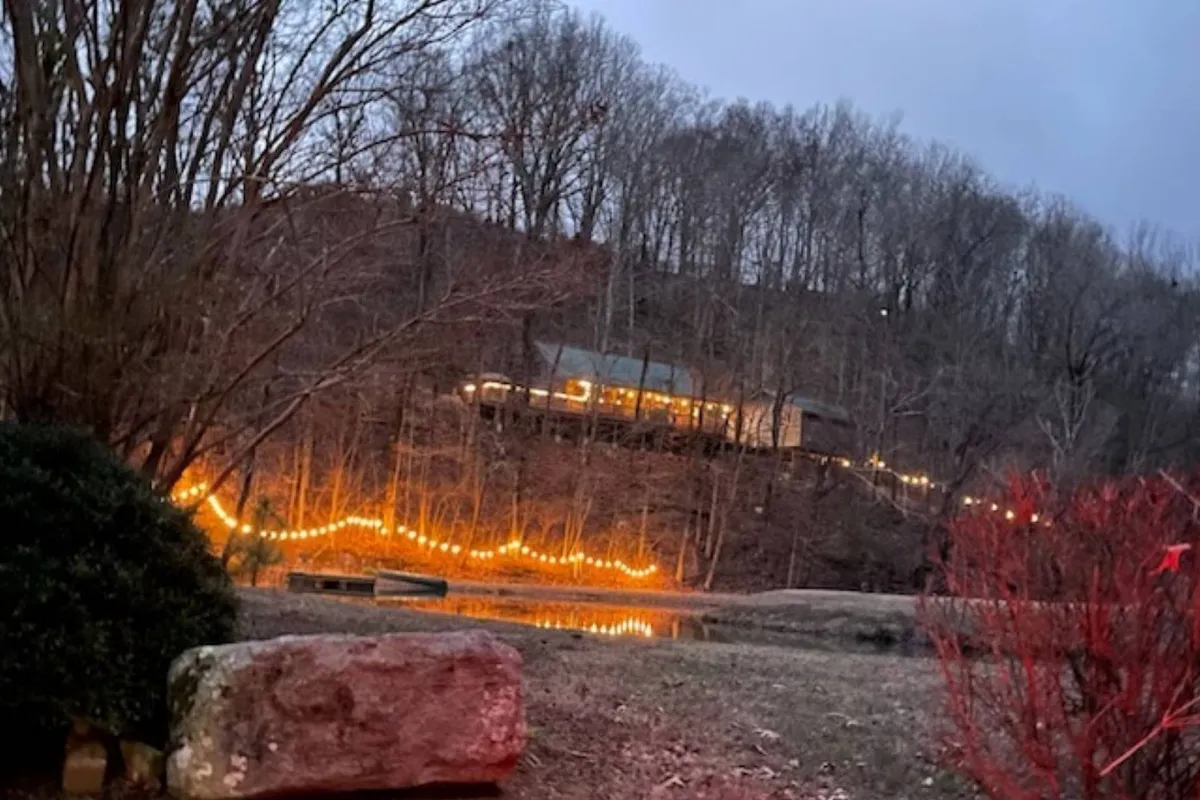 This twilight exterior shot captures the cabin illuminated by twinkling lights, with a reflective water surface and surrounding trees. The scene evokes a sense of evening adventure, tranquil evenings by the water, and the magic of stargazing.