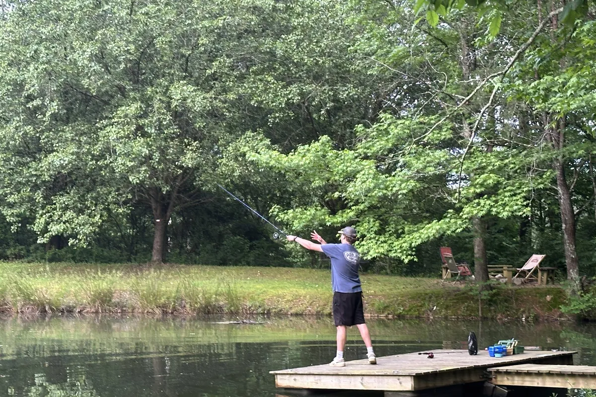 A man fishes from a small dock, lawn chairs set up nearby for relaxing by the water. Trees frame the pond, and the peaceful mood invites guests to enjoy activities like fishing, kayaking, or reflection.