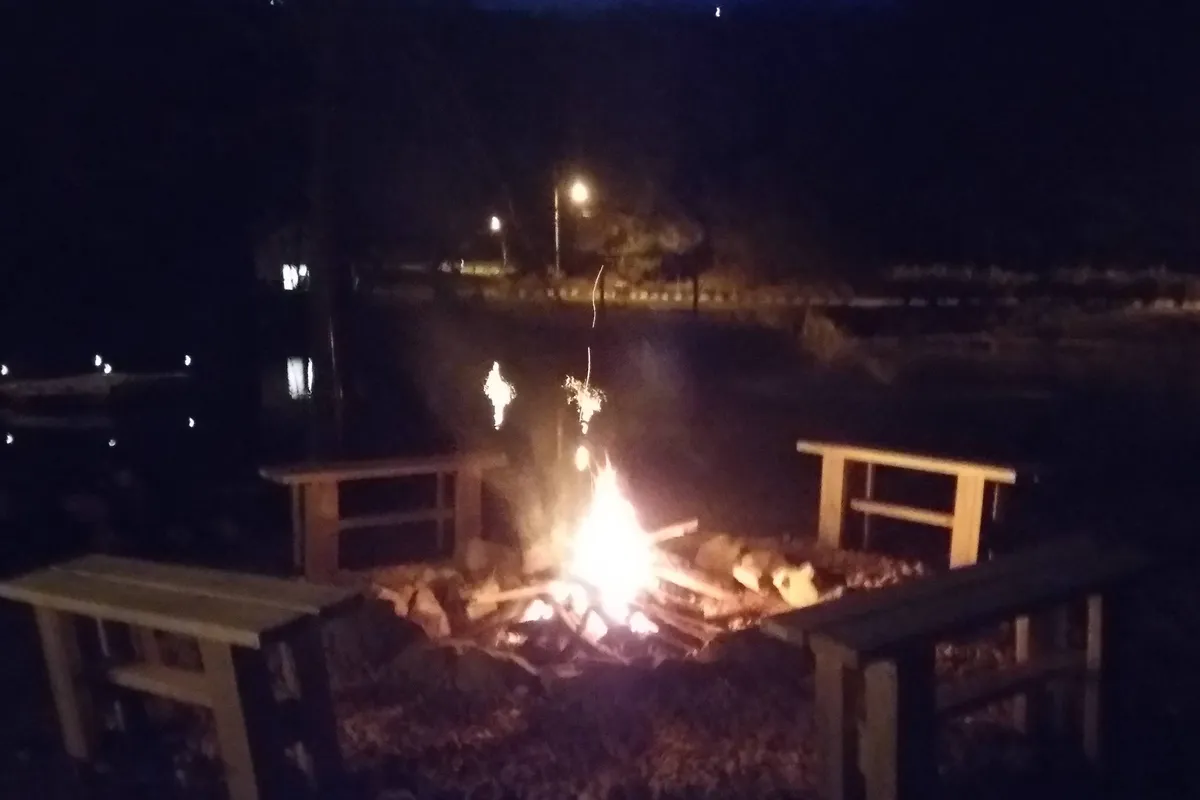 A cozy exterior campfire area featuring wooden benches around a crackling fire, set against the backdrop of a reflective water surface. The ambiance is intimate and relaxed, ideal for evenings spent outdoors.