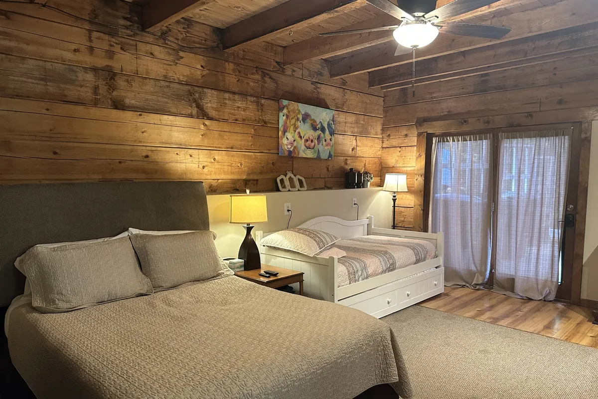 A bedroom featuring a queen-sized bed and a twin bed, set against warm wooden walls. Art on the wall and a bedside lamp add character and comfort, making it an ideal space for families with children or groups seeking flexible sleeping arrangements.
