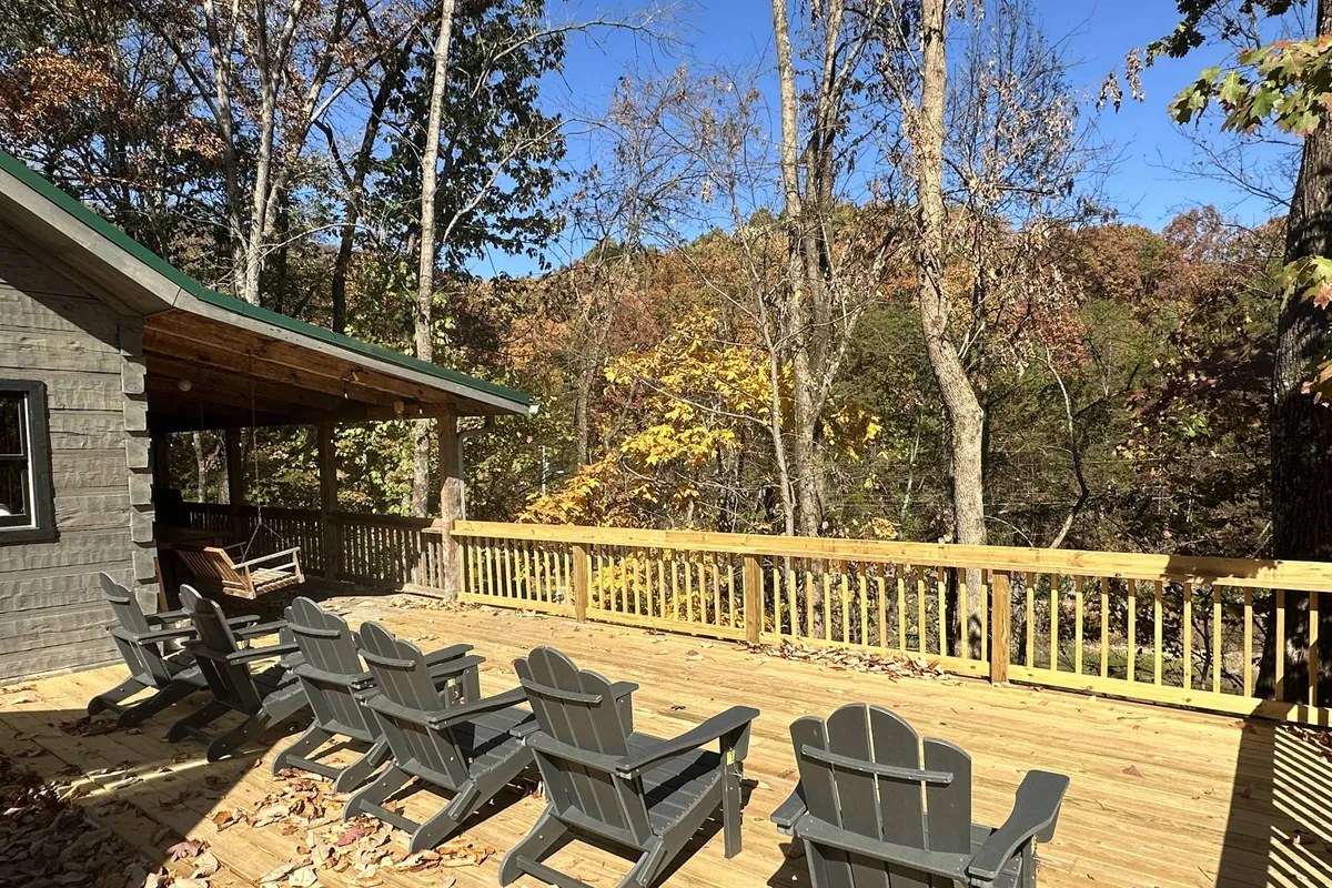 A tranquil exterior shot showing a wooden deck with comfortable chairs and a cabin structure in the background, surrounded by lush trees. The setting is ideal for morning coffee, socializing, or enjoying quiet moments amid nature.
