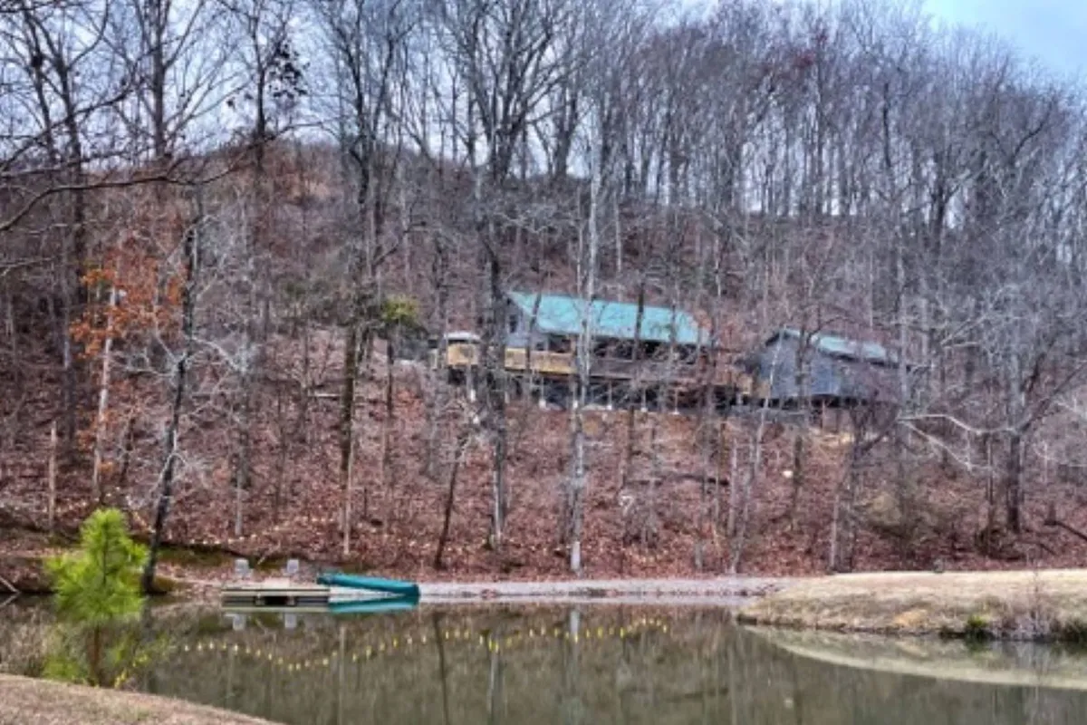 An expansive exterior view shows the house atop a gentle hill overlooking calm water, with a small boat at theshore. The area is surrounded by trees, inviting guests to fish, hike, and take in peaceful nature views.