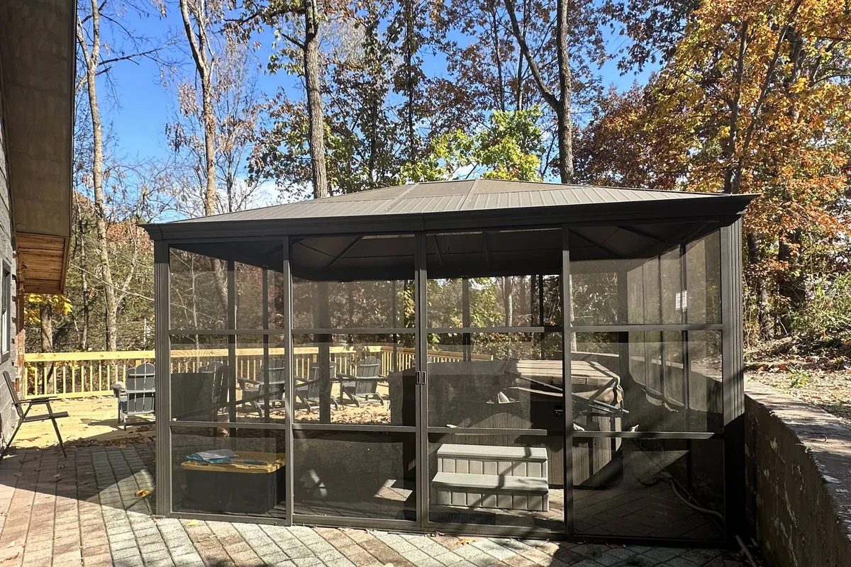 A screened gazebo sits on a wooden deck surrounded by tall trees, offering a sheltered, tranquil space to unwind and enjoy nature regardless of the weather. The area is ideal for relaxation, reading, or enjoying a morning coffee outdoors.
