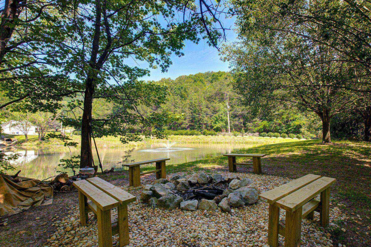 A crackling fire pit is surrounded by benches, set beside a tranquil pond and bordered by tall trees. The arrangement invites gatherings for storytelling, s’mores, or quiet reflection, with fishing and water views adding to the setting’s allure.