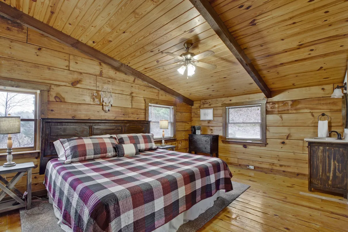 A warmly lit bedroom with a plaid bedspread, wooden walls, and a ceiling fan. The room feels cozy and inviting, perfect for unwinding after a day of family activities. Large windows offer nature views, and soft lamps provide a restful ambiance.