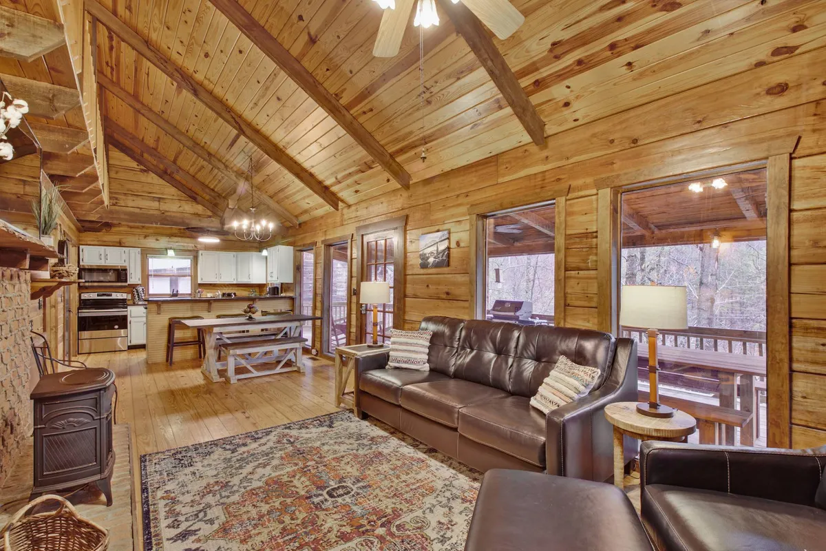 This image showcases a spacious, open-concept living area with visible wooden beams, a cozy sofa, a large rug, a prominent dining table, and the kitchen area in the same view. The scene is filled with warm lighting and inviting textures that encapsulate the communal, log cabin ambiance the section describes.