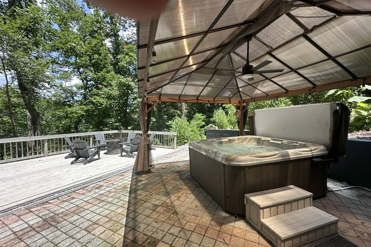 The image features the exterior of a spacious log cabin surrounded by trees, with a steaming hot tub and inviting outdoor seating area set on a large covered deck. The natural setting feels private and serene, perfectly suited for relaxing, socializing, and enjoying the outdoors. The vantage point highlights the connection to nature, with lush greenery and hints of sunset colors peeking through the trees.