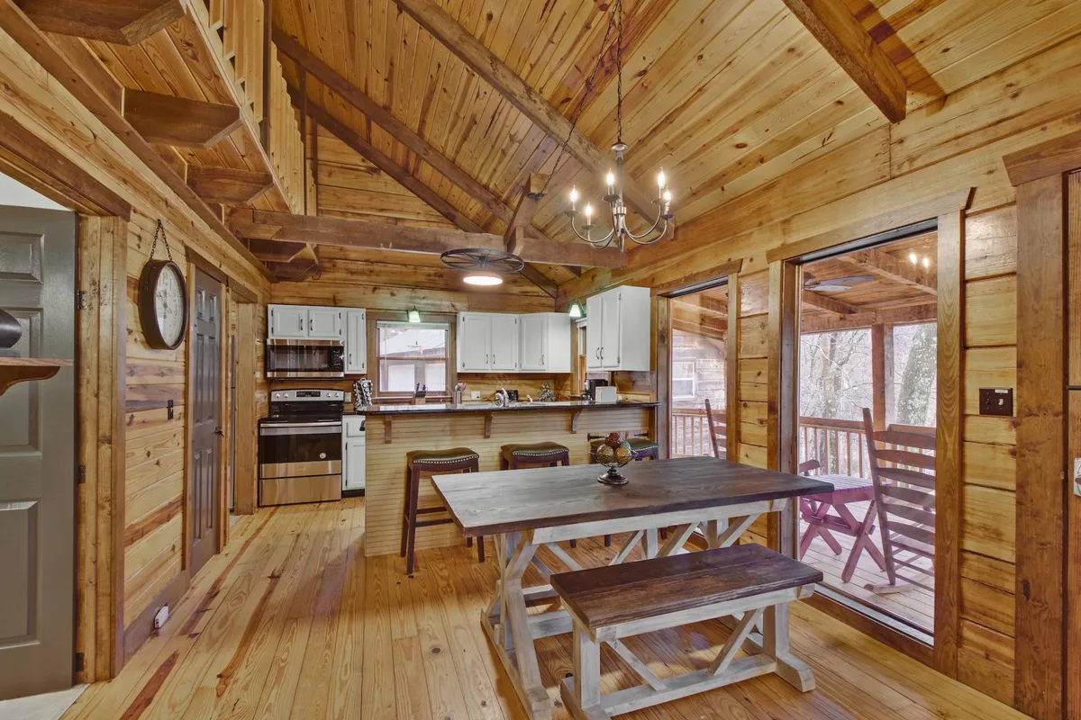 This image focuses on a large dining table next to a kitchen counter, framed by large windows that let in natural light. The ceiling chandelier adds a touch of rustic charm, while the overall mood is intimate and welcoming—ideal for family meals and celebrations.